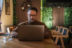 Homem de meia idade com barba, usando óculos, trabalhando em um laptop em um ambiente acolhedor e bem iluminado, com móveis de madeira e plantas ao fundo.