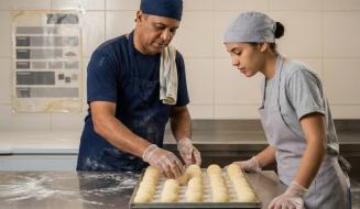 Profissionais de pastelaria/padaria trajando aventais, gorros de proteção e luvas transparentes organizam porções redondas de massa sobre tabuleiro metálico. Na bancada de inox sobressai farinha polvilhada, enquanto ao fundo observa-se parede revestida a azulejos claros com cartaz informativo.