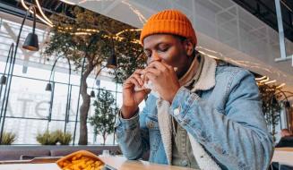 Homem com boné laranja saboreando um sanduíche em um café moderno, cercado por plantas e decoração contemporânea, com batatas fritas ao lado.