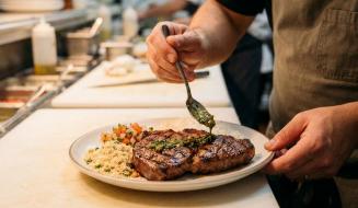 Cozinheiro finaliza a montagem de um prato regando um suculento bife grelhado com molho de ervas. O prato servido na bancada da cozinha também acompanha farofa, vinagrete e arroz.