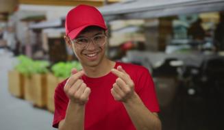 Jovem sorridente usando boné vermelho e óculos transparentes faz gesto com as mãos enquanto está em um ambiente ao ar livre, promovendo estilo casual.