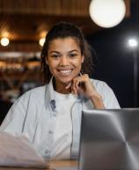 Mulher sorridente sentada à mesa com notebook e segurando papéis em um ambiente de escritório ou café.