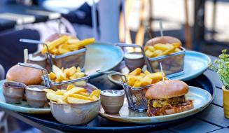 Imagem de uma refeição com batatas fritas e hambúrguer, incluindo molhos e acompanhamentos, servida em um ambiente ao ar livre.