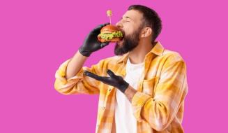 Homem jovem com barba e cabelo curto, usando jaqueta xadrez amarela e branco, segurando um hambúrguer com uma luva preta, pronto para comer, contra um fundo rosa vibrante.