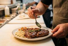 Cozinheiro finaliza a montagem de um prato regando um suculento bife grelhado com molho de ervas. O prato servido na bancada da cozinha também acompanha farofa, vinagrete e arroz.