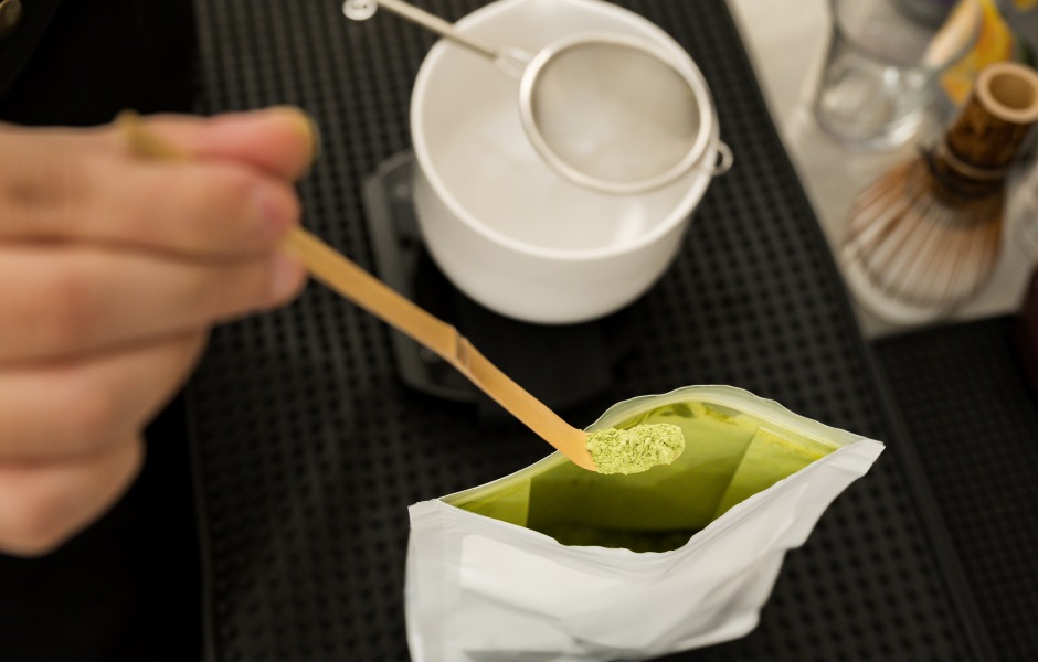 Imagem mostrando a preparação do matcha, com uma colher retirando o pó verde de um pacote, ao lado de uma tigela branca e utensílios para fazer chá.