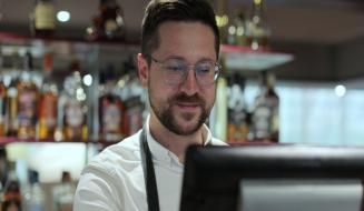Barista profissional atendendo clientes em uma cafeteria, preparado para fazer pedido, com prateleiras de bebidas alcoólicas ao fundo e ambiente moderno.