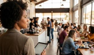 Gerente com cabelo black power segura um tablet e observa com atenção o movimento de um restaurante amplo e iluminado. Ao fundo, clientes ocupam as mesas enquanto a equipe atende no balcão em um ambiente dinâmico e acolhedor.