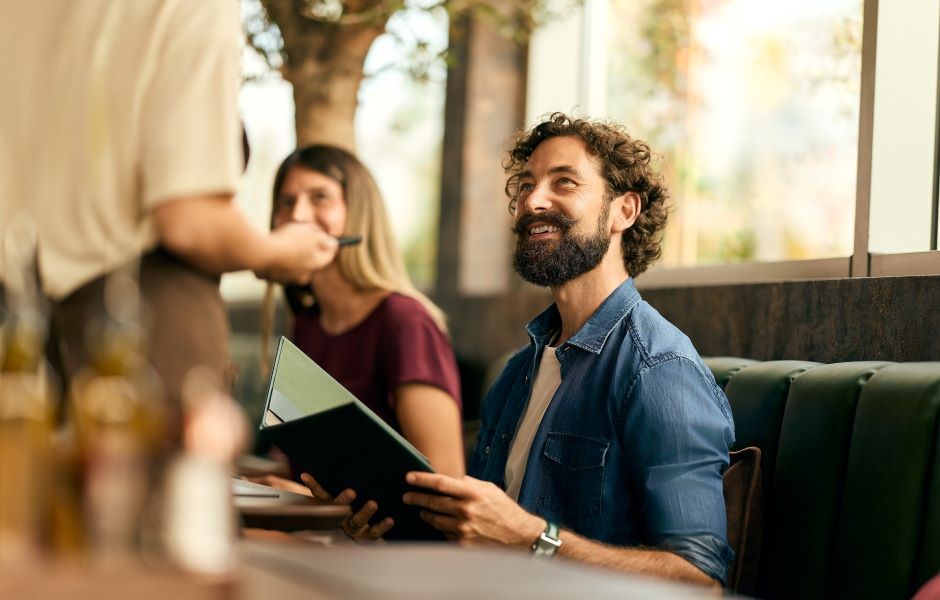 Homem sorridente em um restaurante ou café, interagindo com um garçom enquanto lê um cardápio, ao lado de uma mulher. Ambiente aconchegante com grande janela.