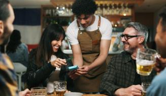 Atendente de avental auxilia clientes em um bar a realizar um pagamento ou pedido via dispositivo móvel.
