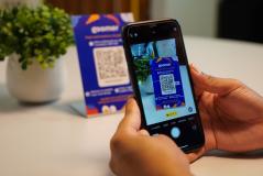 Pessoa escaneando um código QR com um celular na mesa, com uma embalagem azul ao fundo, promovendo a leitura de QR code para acesso a informações e ofertas.
