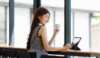 Jovem mulher sorridente utilizando tablet na cafeteria com grande janela de vidro e luz natural, fazendo uma pausa e bebendo chá ou café, promovendo bem-estar e produtividade.
