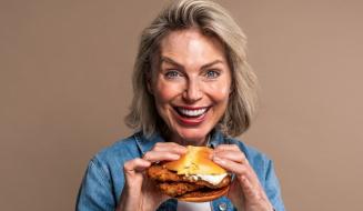 Mulher idosa feliz e sorridente segurando um hambúrguer de frango, destacando a alegria de comer fast food em um fundo neutro.