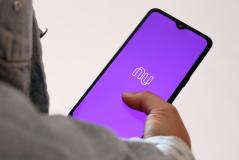 Imagem de um smartphone exibindo o aplicativo Nubank com fundo roxo, mostrando a logo e a interface digital. O usuário interage com a tela, destacando a modernidade e praticidade do aplicativo de serviços financeiros.