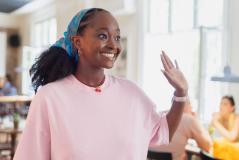 Jovem mulher africana sorridente com cabelo cacheado, usando uma blusa rosa e uma headband azul, em ambiente social, gesticulando com a mão ao lado, transmitindo alegria e entusiasmo.