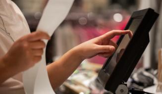 Pessoa operando um sistema de ponto de venda (PDV) em um restaurante, tocando na tela do monitor enquanto segura uma longa nota fiscal de papel.