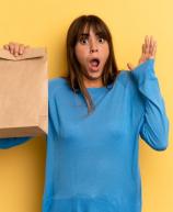 Mulher surpresa segurando sacola de papel com expressão de espanto contra fundo amarelo, representando entrega de comida ou compras online.