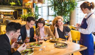 Garçonete de avental azul em pé anota o pedido em um bloco enquanto atende um grupo de quatro jovens sentados ao redor de uma mesa de restaurante com pratos de salada e taças de vinho.