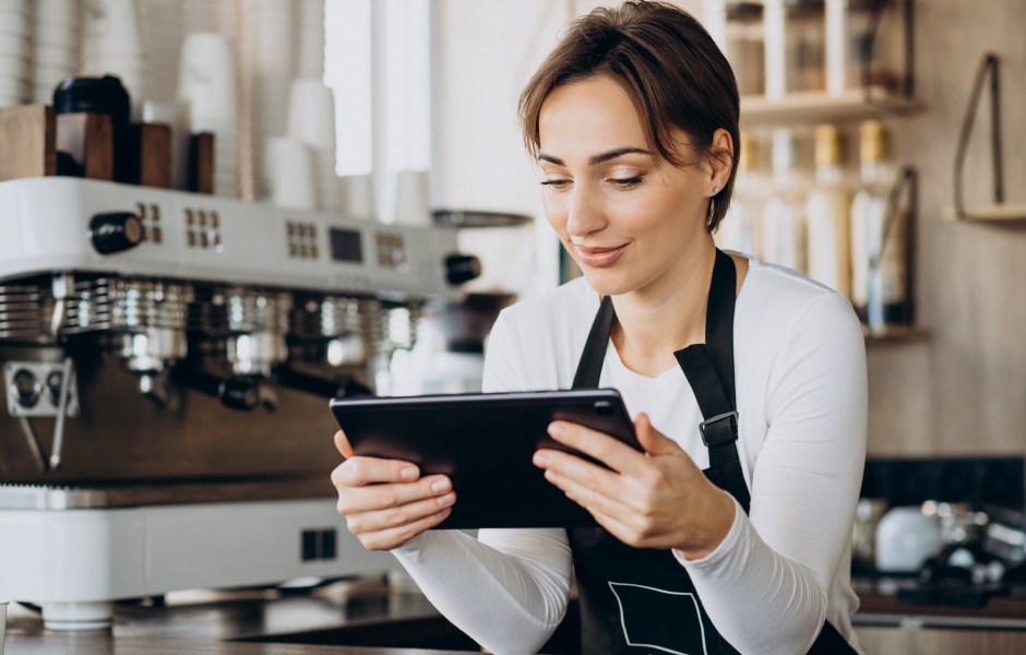 Barista sorridente de avental preto opera um tablet com as duas mãos em um balcão de cafeteria. Ao fundo, destaca-se uma máquina de café expresso profissional branca.