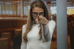 Mulher com óculos apontando para a câmera, destacando-se em um ambiente moderno e acolhedor, ideal para fotos profissionais e retratos.