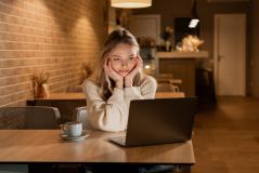 Jovem mulher feliz usando laptop em café aconchegante, focada na tela do computador portátil em um ambiente confortável com iluminação quente.