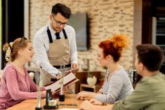 Garçom de avental bege e óculos atende três clientes sentados à mesa de um restaurante, apontando para as opções em um cardápio aberto.